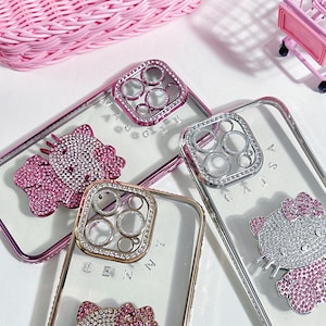 Pu&ograve; includere: Tre custodie per telefono trasparenti con strass e un design Hello Kitty. Le custodie hanno un anello della fotocamera rialzato con strass. Le custodie sono su una superficie bianca.