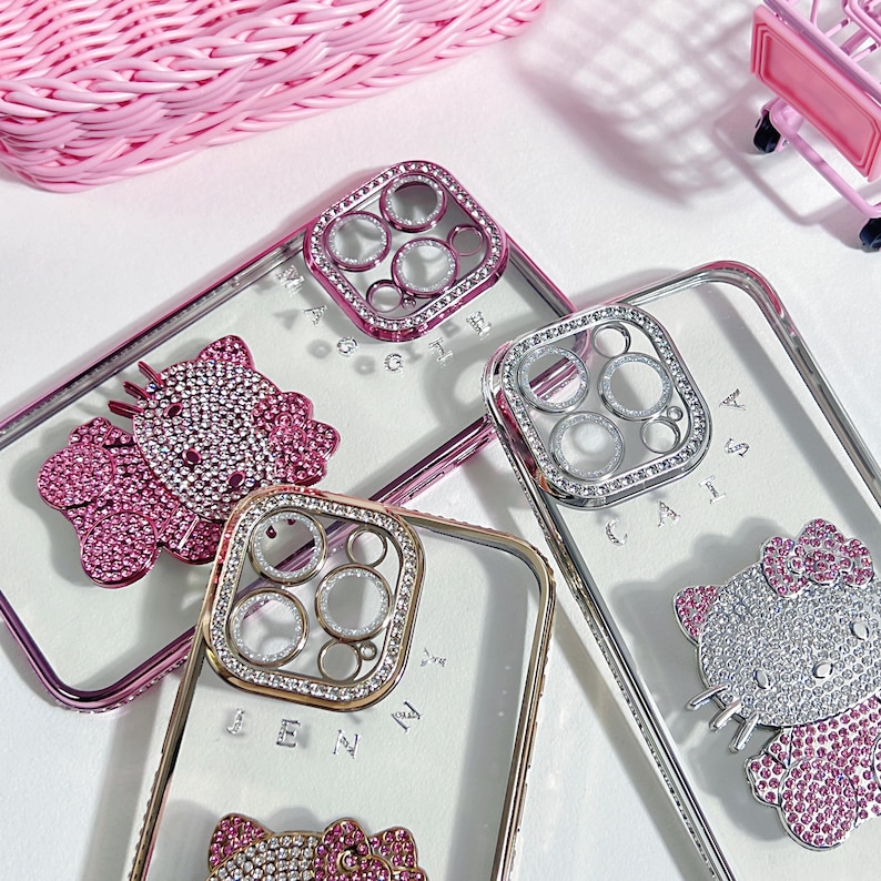 Pu&ograve; includere: Tre custodie per telefono trasparenti con strass e un design Hello Kitty. Le custodie hanno un anello della fotocamera rialzato con strass. Le custodie sono su una superficie bianca.