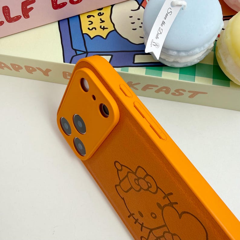 May include: Bright orange phone case with a Hello Kitty design. The case has a smooth finish and a raised camera section. The design is a cartoon outline of Hello Kitty with a bow. The case is on a white surface.