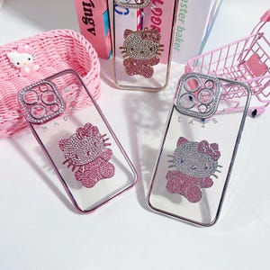 May include: Clear phone cases with a Hello Kitty design encrusted with rhinestones. The cases have a pink or gold frame and a sparkly Hello Kitty figure. The cases are displayed with a pink basket, a pink shopping cart, and books.