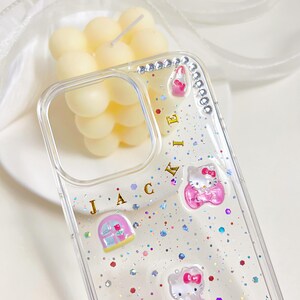 May include: Clear phone case with glitter and Hello Kitty embellishments. The case features a decorative design with pink and white Hello Kitty figures, rhinestones, and the letters J, A, C, E, I, K. A decorative candle is in the background.