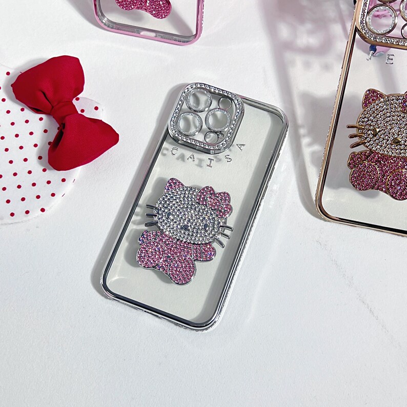K&ouml;nnte beinhalten: Transparente Handyh&uuml;lle mit silbernem Rahmen und einem Strass-Hello Kitty-Design. Die Hello Kitty-Figur besteht aus silbernen und rosa Strasssteinen. Eine rote Schleife und ein wei&szlig;es Stoff mit Polka-Dots befinden sich im Hintergrund.