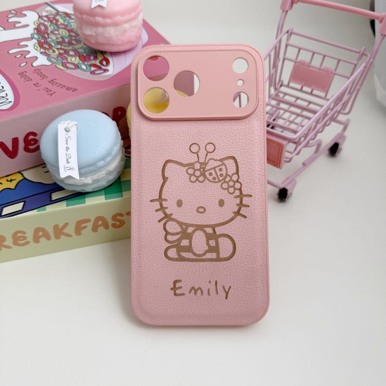 Pu&ograve; includere: Custodia per telefono rosa con un design di Hello Kitty e il nome "Emily" in corsivo. La custodia ha un ritaglio per la fotocamera e una superficie strutturata. Un carrello della spesa giocattolo rosa &egrave; sullo sfondo.