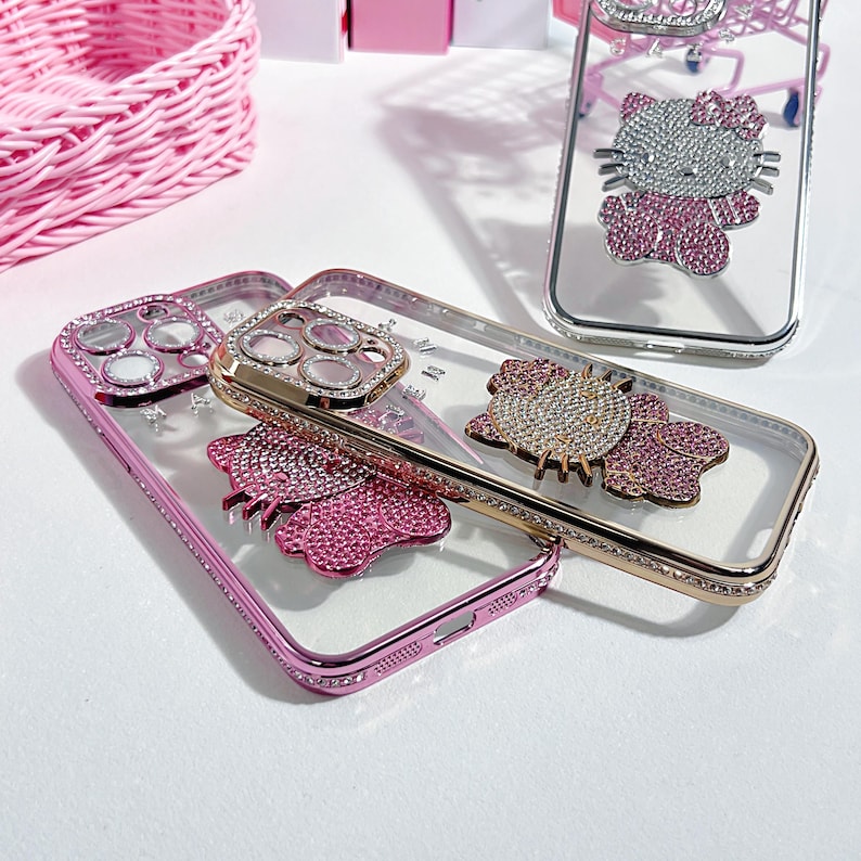 May include: Three clear phone cases with pink and silver rhinestones. The cases feature a butterfly, a Hello Kitty character, and a camera lens cover.
