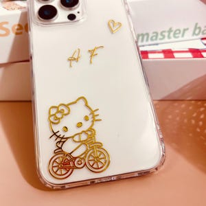 Op de afbeelding: Een witte iPhone-hoes met een gouden Hello Kitty-ontwerp. Het ontwerp toont Hello Kitty op een fiets. De hoes heeft ook de letters "AF" en een hart in goud.