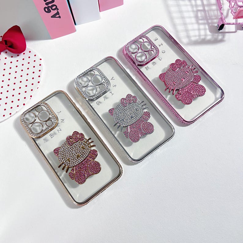 K&ouml;nnte beinhalten: Drei transparente Handyh&uuml;llen mit einem Hello-Kitty-Design. Jede H&uuml;lle hat einen anderen Farbrahmen: rosa, silber und gold. Die H&uuml;llen haben eine transparente R&uuml;ckseite und ein Hello-Kitty-Design mit Strasssteinen.