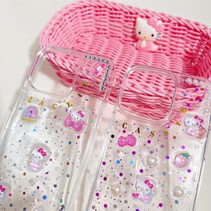 May include: Clear phone cases with glitter and Hello Kitty embellishments. The cases feature various Hello Kitty designs, small charms, and the word "JACKY" and "CAS" in gold letters. A pink woven basket and a Hello Kitty figurine are in the background.