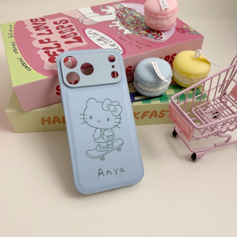 May include: A light blue phone case featuring a Hello Kitty design on a skateboard, with the name "Anya" below. The case has a camera cutout at the top. The image also shows decorative items like macaron-shaped candles and a miniature shopping cart.