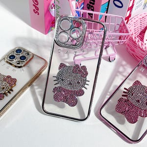 K&ouml;nnte beinhalten: Drei transparente Handyh&uuml;llen mit Strasssteinen. Die H&uuml;llen zeigen ein Hello Kitty-Design mit rosa und silbernen Strasssteinen. Die H&uuml;llen sind f&uuml;r ein iPhone mit einer Kamerabuckel.