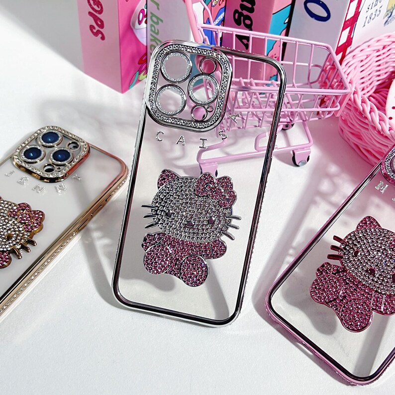 K&ouml;nnte beinhalten: Drei transparente Handyh&uuml;llen mit Strasssteinen. Die H&uuml;llen zeigen ein Hello Kitty-Design mit rosa und silbernen Strasssteinen. Die H&uuml;llen sind f&uuml;r ein iPhone mit einer Kamerabuckel.