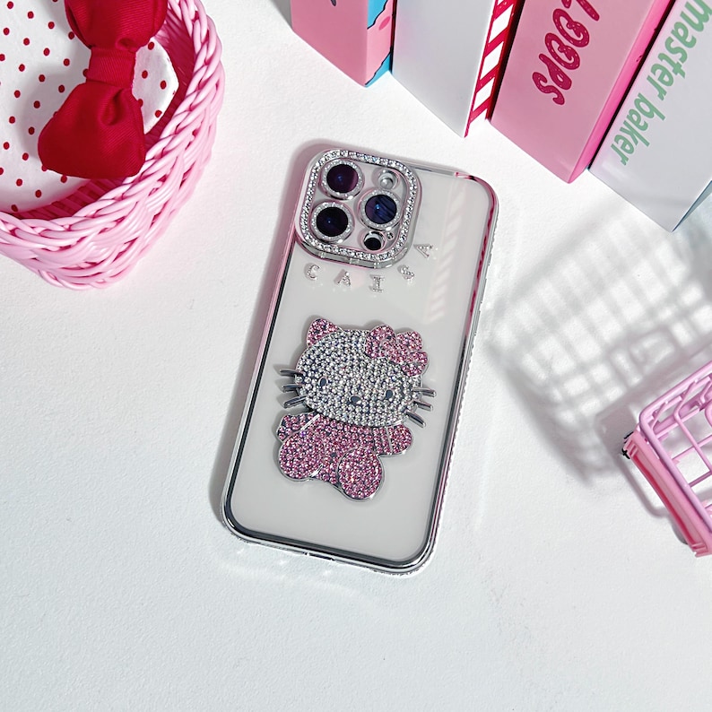 May include: Clear phone case with a silver frame and a sparkling Hello Kitty design. The case features a pink bow and a rhinestone-studded cat. The word "CAI" is visible on the case.