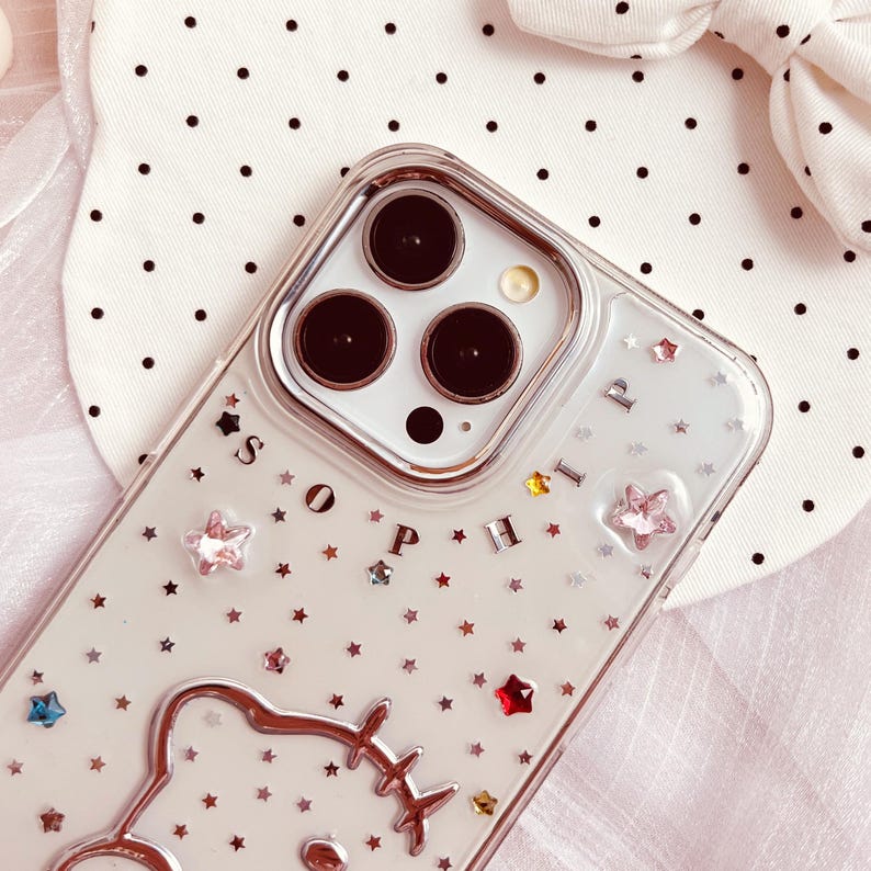 May include: A clear phone case with a silver glitter background and a silver bear outline. The case has a silver inscription that reads 'SOPHIA' and features a variety of colorful rhinestones. The phone case is designed for a phone with a triple lens camera system.