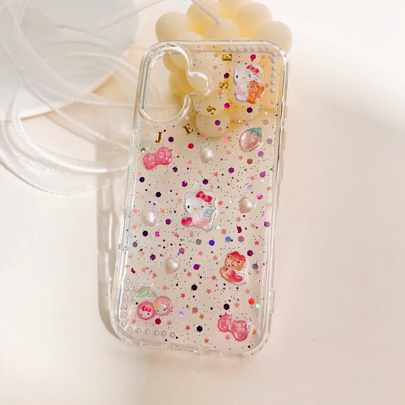 Peut inclure: Coque de t&eacute;l&eacute;phone transparente avec un design de paillettes rose, blanc et or. La coque pr&eacute;sente des personnages Hello Kitty, des cerises et des &eacute;toiles. La coque porte &eacute;galement les lettres "J E S S E".