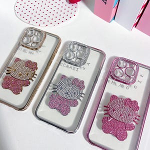 May include: Three clear phone cases with rhinestones and a Hello Kitty design. The cases are decorated with rhinestones and have a clear back. The cases are labeled "Queen", "Caissa", and "Magoo".