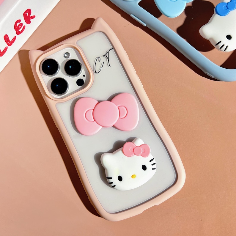 Puede incluir: Una funda de tel&eacute;fono rosa y blanca con la parte posterior transparente. La funda tiene un lazo rosa y un personaje de Hello Kitty blanco con un lazo rosa en la parte superior.