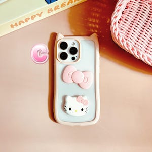 Puede incluir: Una funda de tel&eacute;fono rosa y blanca con un dise&ntilde;o de Hello Kitty. La funda tiene una parte posterior transparente y un lazo rosa en la parte posterior. La funda est&aacute; dise&ntilde;ada para un iPhone.