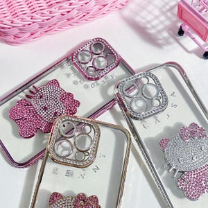 K&ouml;nnte beinhalten: Drei transparente Handyh&uuml;llen mit Strasssteinen und einem Hello-Kitty-Design. Die H&uuml;llen haben einen erh&ouml;hten Kameraring mit Strasssteinen. Die H&uuml;llen liegen auf einer wei&szlig;en Oberfl&auml;che.