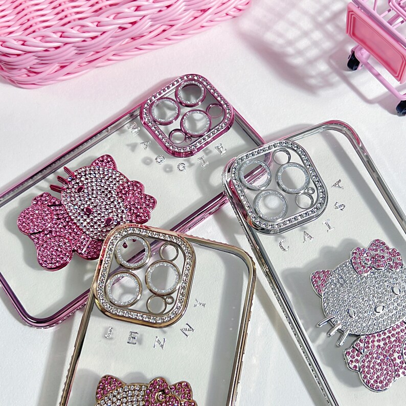 K&ouml;nnte beinhalten: Drei transparente Handyh&uuml;llen mit Strasssteinen und einem Hello-Kitty-Design. Die H&uuml;llen haben einen erh&ouml;hten Kameraring mit Strasssteinen. Die H&uuml;llen liegen auf einer wei&szlig;en Oberfl&auml;che.