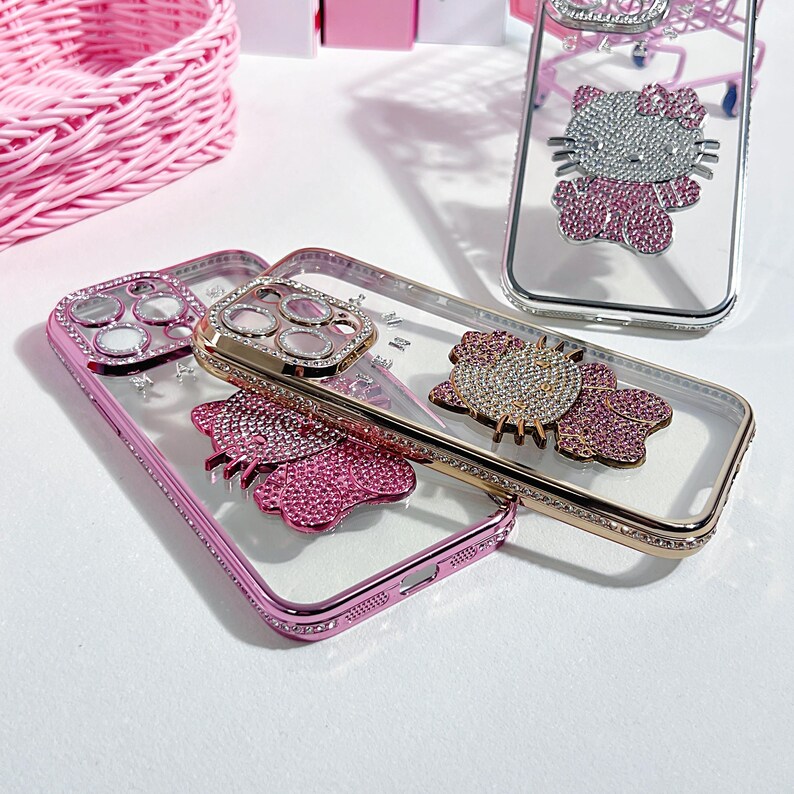 May include: Clear phone cases with pink and gold metallic frames. Each case features a bedazzled design, including a bow and a character. The cases are embellished with rhinestones and are displayed on a white surface.