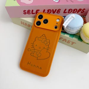 Puede incluir: Funda de tel&eacute;fono naranja con un dise&ntilde;o de Hello Kitty y el nombre "Hanna" grabado. La funda tiene un recorte para la c&aacute;mara. La funda est&aacute; junto a una caja con las palabras "SELF LOVE LOOPS".