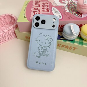 Puede incluir: Una funda de tel&eacute;fono azul claro con un dise&ntilde;o de Hello Kitty en monopat&iacute;n. La funda tiene un m&oacute;dulo de c&aacute;mara azul claro y el nombre "Anya" impreso debajo. La funda est&aacute; sobre una mesa con una cesta rosa y una caja.