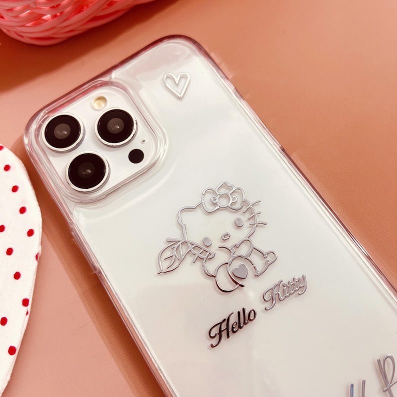 May include: Clear phone case with a silver Hello Kitty design. The case features a raised silver Hello Kitty figure holding a heart, a silver heart, and the words "Hello Kitty" in cursive.