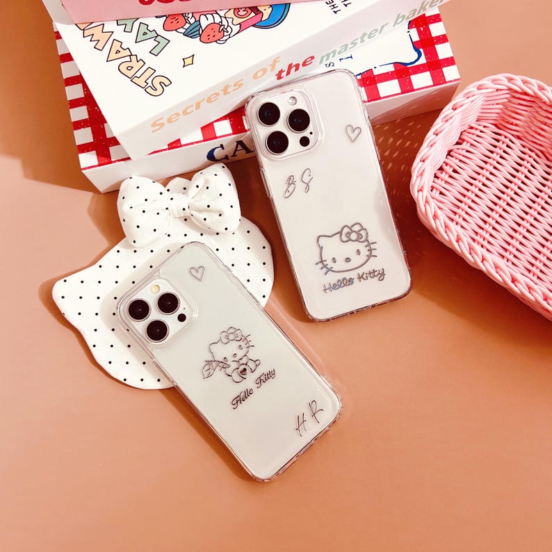 Op de afbeelding: Twee transparante telefoonhoesjes met Hello Kitty-ontwerpen. De hoesjes zijn transparant en laten de camera van de telefoon zien. Een hoesje heeft een Hello Kitty met een strik, de andere heeft een Hello Kitty met een bloem.