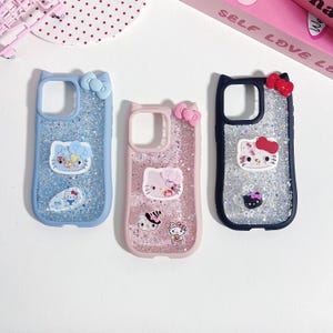 Op de afbeelding: Drie telefoonhoesjes met Hello Kitty-ontwerpen en glitter. De hoesjes zijn lichtblauw, roze en marineblauw, elk met een strik aan de bovenkant. De Hello Kitty-graphics bevatten ballonnen, sushi en andere schattige elementen.