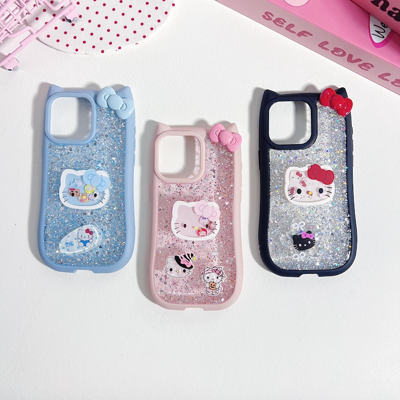 Op de afbeelding: Drie telefoonhoesjes met Hello Kitty-ontwerpen en glitter. De hoesjes zijn lichtblauw, roze en marineblauw, elk met een strik aan de bovenkant. De Hello Kitty-graphics bevatten ballonnen, sushi en andere schattige elementen.