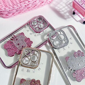 K&ouml;nnte beinhalten: Drei transparente Handyh&uuml;llen mit Strasssteinen und einem Hello-Kitty-Design. Die H&uuml;llen haben einen erh&ouml;hten Kameraring mit Strasssteinen. Die H&uuml;llen liegen auf einer wei&szlig;en Oberfl&auml;che.