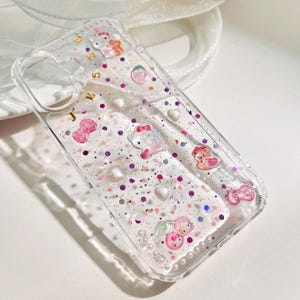 Peut inclure: Coque de t&eacute;l&eacute;phone transparente avec un design de paillettes rose, blanc et violet. La coque pr&eacute;sente divers personnages de dessins anim&eacute;s, dont Hello Kitty, un ours et une fraise.
