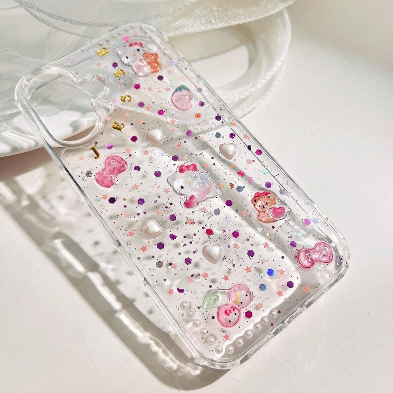Peut inclure: Coque de t&eacute;l&eacute;phone transparente avec un design de paillettes rose, blanc et violet. La coque pr&eacute;sente divers personnages de dessins anim&eacute;s, dont Hello Kitty, un ours et une fraise.