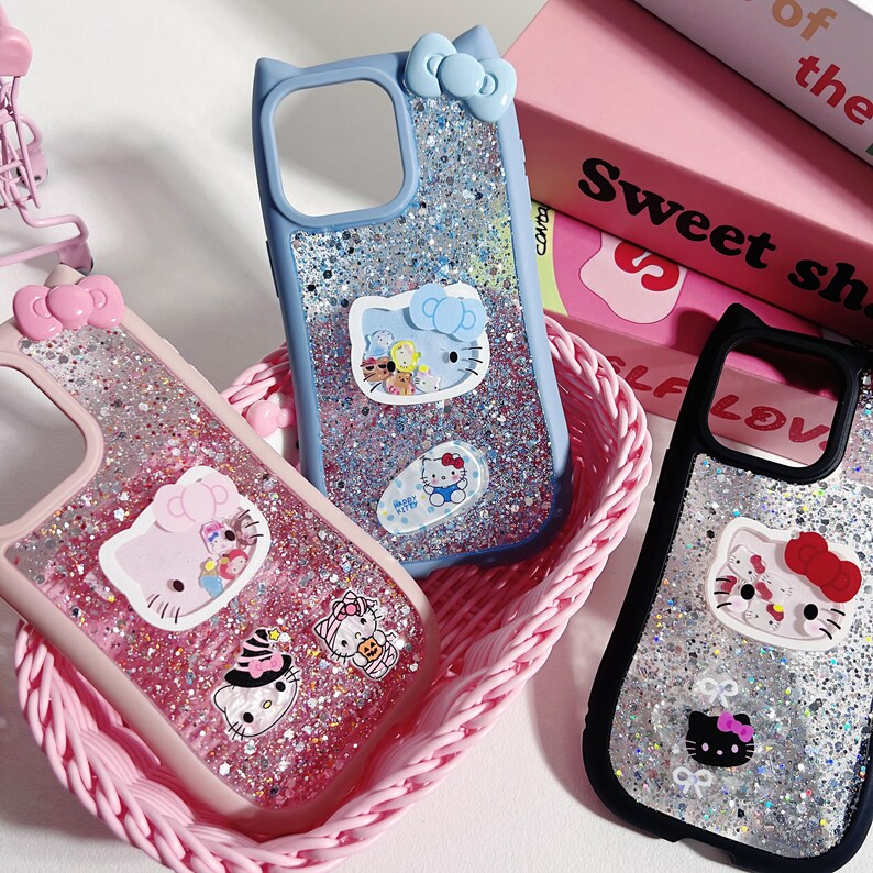 Op de afbeelding: Een verzameling glitterende telefoonhoesjes met Hello Kitty-ontwerpen. De hoesjes zijn roze, blauw en zwart, met verschillende Hello Kitty-stickers. De hoesjes liggen in een roze geweven mand, met een roze boek op de achtergrond.