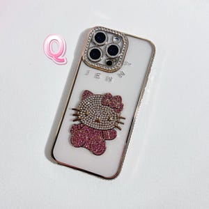 May include: White phone case with a gold frame and a Hello Kitty design. The Hello Kitty is embellished with pink rhinestones. The camera area is surrounded by a gold frame with clear rhinestones. The word "JENNY" is printed on the case.