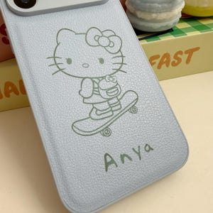 Puede incluir: Funda de tel&eacute;fono azul claro con una ilustraci&oacute;n de Hello Kitty en monopat&iacute;n. El nombre "Anya" est&aacute; impreso debajo. La funda tiene una superficie texturizada y est&aacute; dise&ntilde;ada para proteger el tel&eacute;fono.