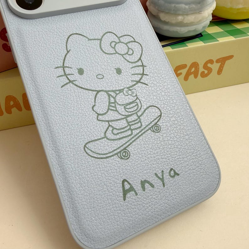 Puede incluir: Funda de tel&eacute;fono azul claro con una ilustraci&oacute;n de Hello Kitty en monopat&iacute;n. El nombre "Anya" est&aacute; impreso debajo. La funda tiene una superficie texturizada y est&aacute; dise&ntilde;ada para proteger el tel&eacute;fono.