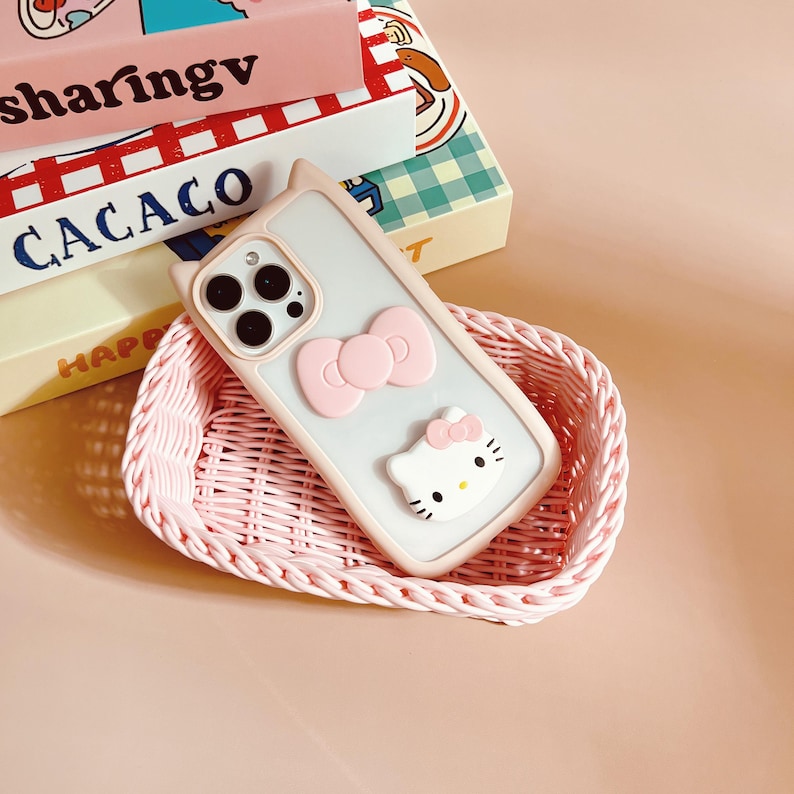 以下が含まれることがあります： ピンクと白の携帯電話ケースで、ハローキティのデザインとピンクのリボンが付いています。ケースはピンクの籐のバスケットに入っています。
