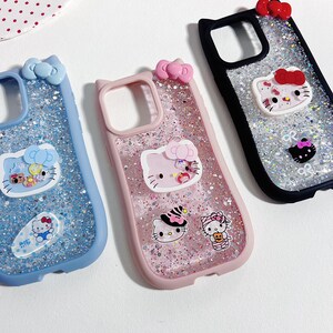 Op de afbeelding: Drie telefoonhoesjes met glitter en Hello Kitty-ontwerpen. E&eacute;n hoesje is blauw, &eacute;&eacute;n roze en &eacute;&eacute;n zwart. Elk hoesje heeft een strik aan de bovenkant en verschillende Hello Kitty-personagestickers.