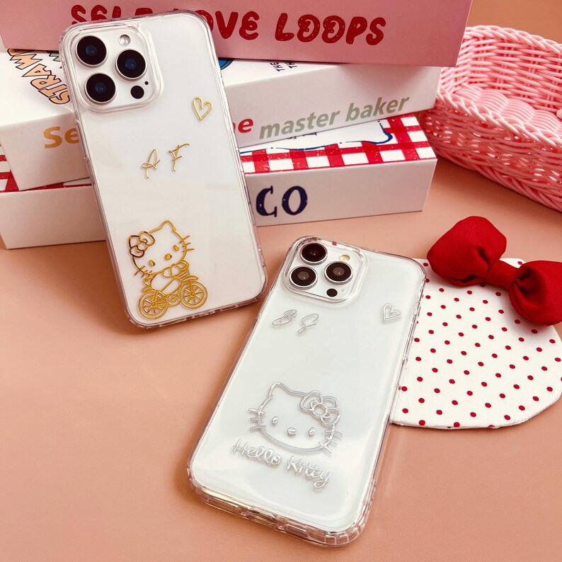 Op de afbeelding: Twee transparante telefoonhoesjes met Hello Kitty-ontwerpen. Een hoesje heeft een gouden Hello Kitty-ontwerp en de andere een zilveren Hello Kitty-ontwerp. Beide hoesjes hebben een transparante achtergrond.