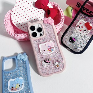 Może przedstawiać: R&oacute;żowe, niebieskie i czarne etui na telefony z brokatem i wzorami Hello Kitty. R&oacute;żowe etui ma kokardkę i kilka naklejek Hello Kitty. Niebieskie etui ma kokardkę i wz&oacute;r Hello Kitty. Czarne etui ma wz&oacute;r Hello Kitty.