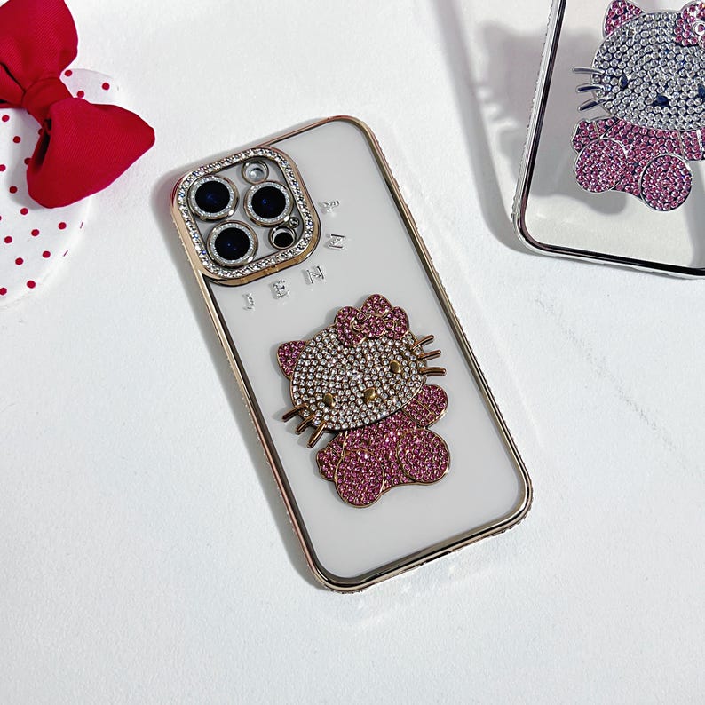 K&ouml;nnte beinhalten: Eine wei&szlig;e iPhone-H&uuml;lle mit einem rosafarbenen Hello-Kitty-Design und Strasssteinen. Die H&uuml;lle hat eine transparente Kameraobjektivabdeckung mit goldfarbenem Rand.