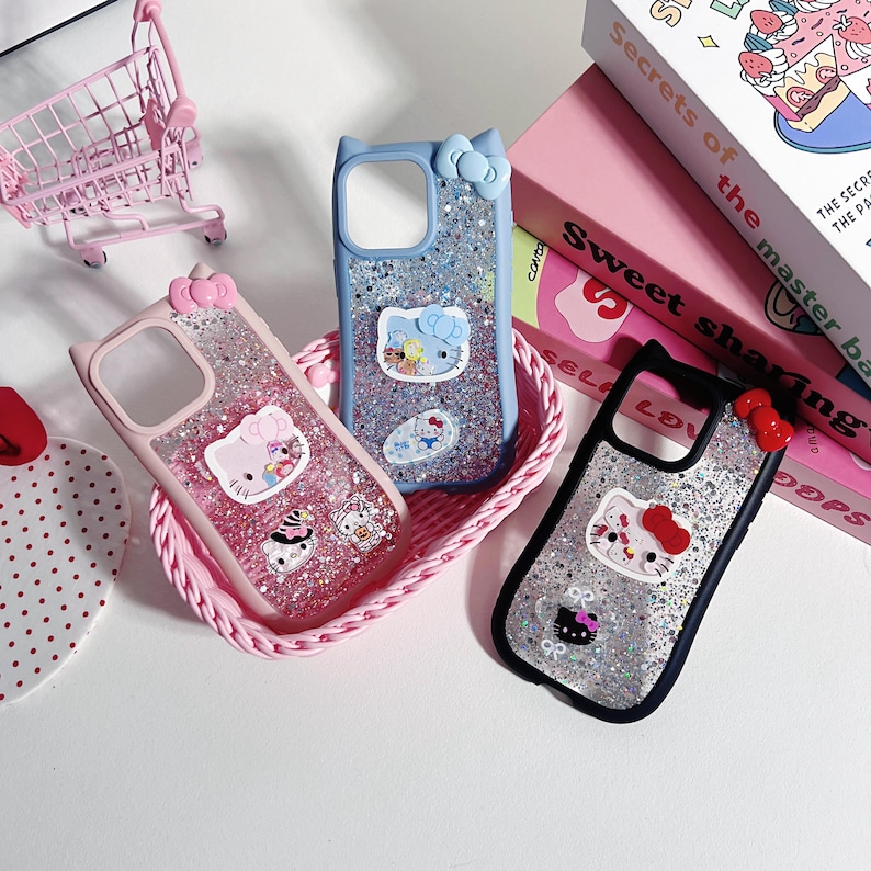 Op de afbeelding: Drie telefoonhoesjes met Hello Kitty-ontwerpen en glitter. Een roze, een blauwe en een zwarte hoes worden gepresenteerd in een roze mand. Een roze winkelwagentje is zichtbaar op de achtergrond.