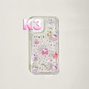 May include: Clear phone case with a glittery design and Hello Kitty character embellishments. The case features a pink "K3" graphic and the word "SHE" in gold lettering. Additional decorative elements include a rainbow and small pink hearts.