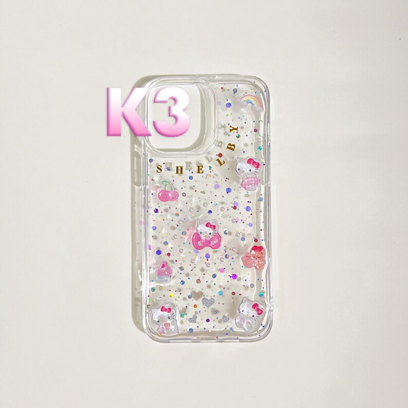 May include: Clear phone case with a glittery design and Hello Kitty character embellishments. The case features a pink "K3" graphic and the word "SHE" in gold lettering. Additional decorative elements include a rainbow and small pink hearts.