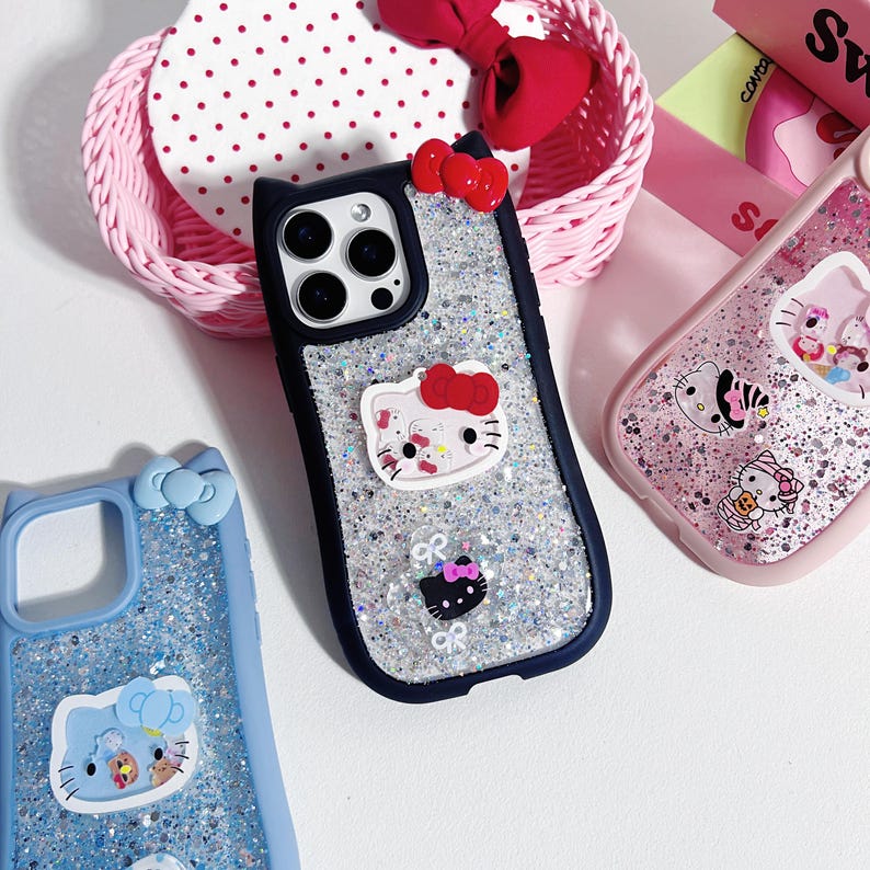 Op de afbeelding: Een verzameling telefoonhoesjes met Hello Kitty-ontwerpen. De hoesjes zijn in verschillende kleuren, waaronder blauw, roze en zilver, met glitter- en strikaccenten. E&eacute;n hoesje heeft een zwarte rand en een 3D Hello Kitty-figuur.