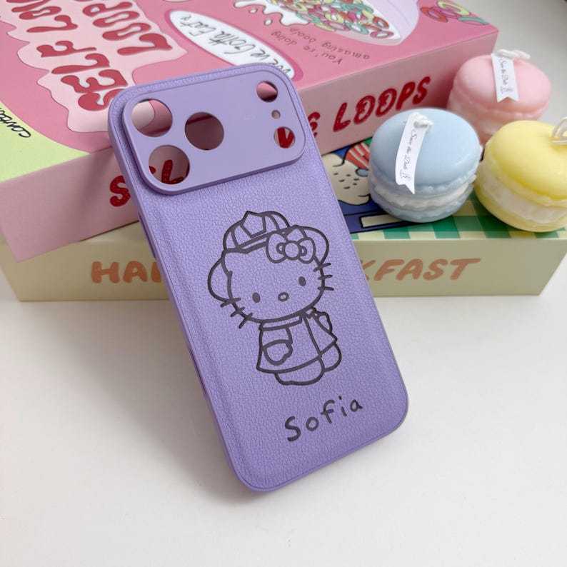 Puede incluir: Una funda de tel&eacute;fono lavanda con un dise&ntilde;o de Hello Kitty y el nombre "Sofia" impreso debajo. La funda tiene una apariencia texturizada y un protector de lente de c&aacute;mara. El fondo incluye una caja de cereales rosa y macarons decorativos.