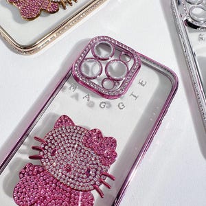May include: A clear phone case with a pink Hello Kitty design and a pink jeweled camera lens cover. The case has the word "MAGGIE" written on it.