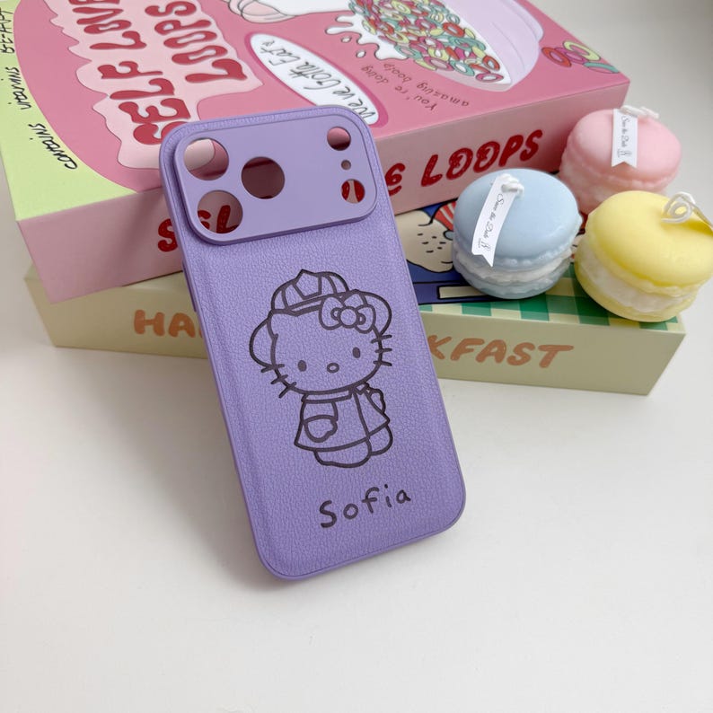 Pu&ograve; includere: Una custodia per telefono lilla con un design di Hello Kitty e il nome "Sofia" stampato sotto. La custodia ha un ritaglio per la fotocamera ed &egrave; accanto a una scatola di cereali e macaron decorativi.