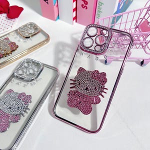 K&ouml;nnte beinhalten: Drei transparente Handyh&uuml;llen mit Strasssteinen und einem Hello-Kitty-Design. Die H&uuml;llen sind rosa, silber und gold. Das Hello-Kitty-Design ist rosa mit Strasssteinen.