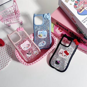 Może przedstawiać: Trzy etui na telefon z motywami Hello Kitty i brokatem. R&oacute;żowe, niebieskie i czarne etui są prezentowane w r&oacute;żowym koszyku. R&oacute;żowy w&oacute;zek na zakupy jest widoczny w tle.